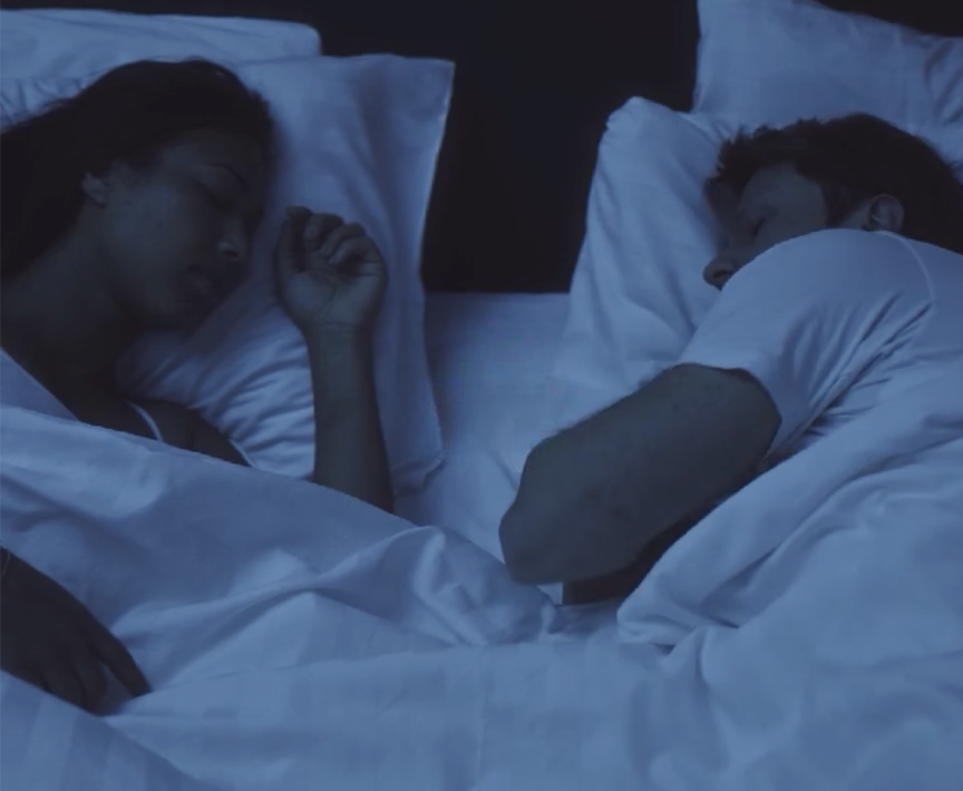 Een vrouw en een man liggen tegenover elkaar in bed te slapen onder witte lakens en kussens, verlicht door zacht blauw licht.