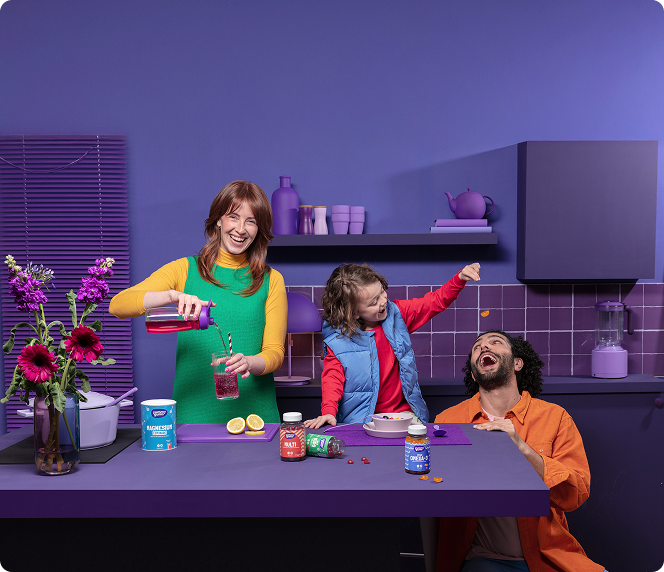 Een lachende vrouw schenkt een drankje in terwijl een kind een lachende man vitaminen voert in een kleurrijke paarse keuken met bloemen en potten op het aanrecht.