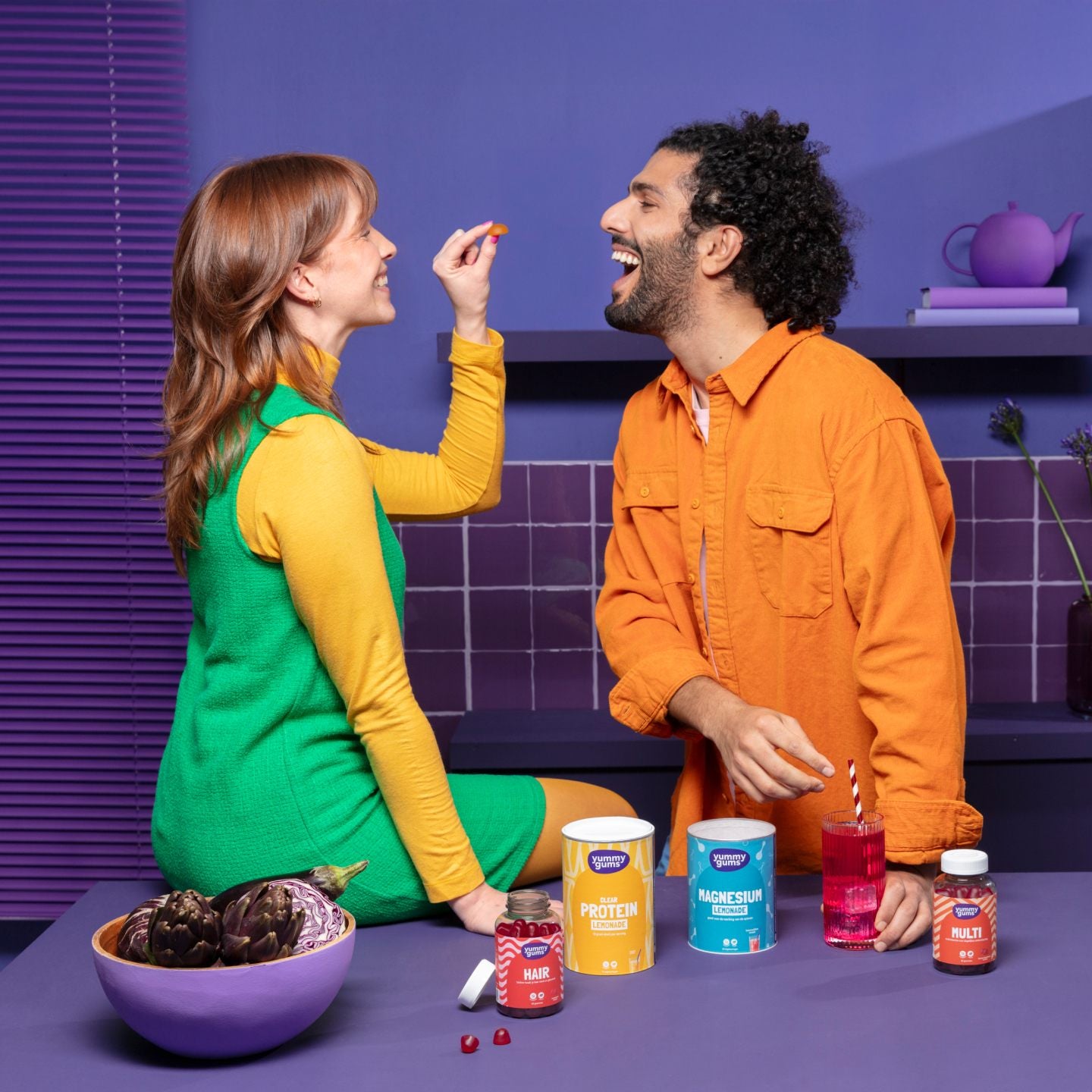 Een vrouw in een groene jurk en geel topje voert een gummy aan een lachende man in een oranje shirt. Ze worden omringd door kleurrijke supplementencontainers op een paars aanrecht met artisjokken.
